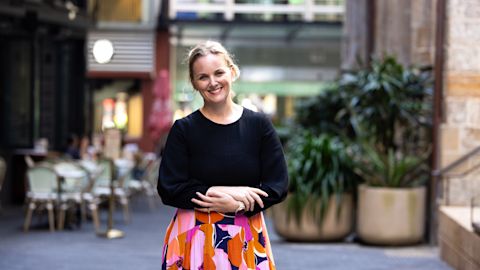 Woman in a black top and colorful skirt standing in an outdoor area with cafes and plants in the background.