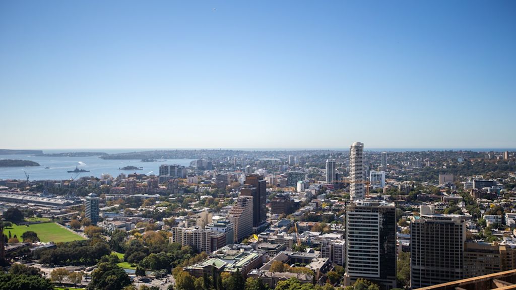 Cityscape view of Sydney, showing skyscrapers, green spaces, and water in the background under a clear blue sky.