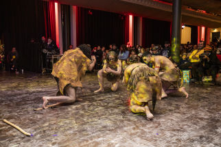 Four Aboriginal dancers crouch low in a circle as they perform for an audience indoors