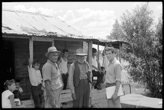 A group of men and children stand in front of a wooden house with a tin roof. Children and young people lean, sit and stand around outside the house.