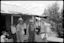 A group of men and children stand in front of a wooden house with a tin roof. Children and young people lean, sit and stand around outside the house.