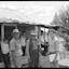 A group of men and children stand in front of a wooden house with a tin roof. Children and young people lean, sit and stand around outside the house.