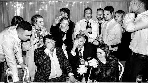 A group of people in formal attire, standing and sitting around a table, appear to be in shock or disbelief, with some covering their faces.