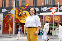 A woman in a white shirt and yellow pants stands on a city street with a dragon mural in the background.