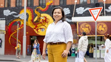 A woman in a white shirt and yellow pants stands on a city street with a dragon mural in the background.