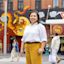 A woman in a white shirt and yellow pants stands on a city street with a dragon mural in the background.