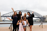 Five people and two children smile and wave in front of the Sydney Harbour Bridge.
