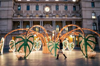 A woman walks past a large outdoor art installation featuring green, leaf-shaped structures and bamboo arches in front of a historic building with a clock.