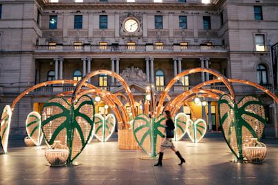 A woman walks past a large outdoor art installation featuring green, leaf-shaped structures and bamboo arches in front of a historic building with a clock.