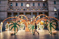 A woman walks past a large outdoor art installation featuring green, leaf-shaped structures and bamboo arches in front of a historic building with a clock.