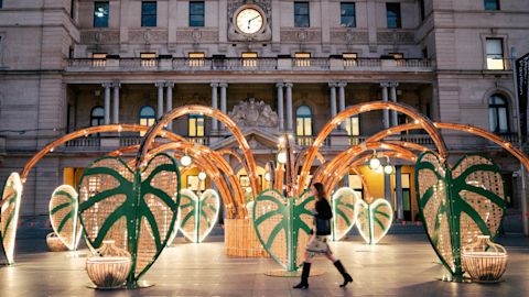 A woman walks past a large outdoor art installation featuring green, leaf-shaped structures and bamboo arches in front of a historic building with a clock.