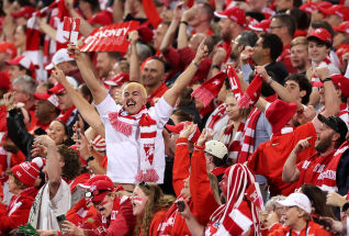 A crowd of excited Sydney Swans fans dressed in red and white cheer passionately at a stadium, with one fan raising a broken flag in celebration.