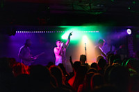 A band performs on stage under colorful lights in front of a crowd at a concert venue. The lead singer gestures with one arm raised.