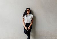 A woman with long dark hair, wearing a sleeveless white top and black pants, stands smiling against a plain light gray wall.