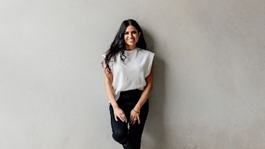 A woman with long dark hair, wearing a sleeveless white top and black pants, stands smiling against a plain light gray wall.