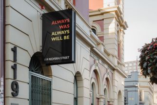 A black banner with the text "Always Was Always Will Be" hangs on a building facade. Mounted on the wall are the numbers 119. 