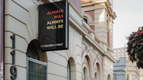 A black banner with the text "Always Was Always Will Be" hangs on a building facade. Mounted on the wall are the numbers 119.