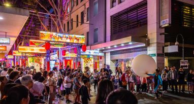 A large crowd gathers on a city street adorned with colorful decorations and lights. A person with a large balloon stands among the crowd. Some signs and a building with balconies are visible.