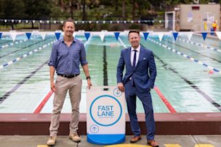 Two men stand beside a “Fast Lane” swim sign at an outdoor pool, one wearing a blue shirt and khakis, the other in a blue suit and tie.