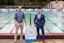Two men stand beside a “Fast Lane” swim sign at an outdoor pool, one wearing a blue shirt and khakis, the other in a blue suit and tie.