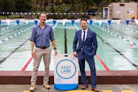 Two men stand beside a “Fast Lane” swim sign at an outdoor pool, one wearing a blue shirt and khakis, the other in a blue suit and tie.