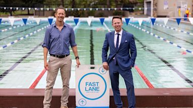 Two men stand beside a “Fast Lane” swim sign at an outdoor pool, one wearing a blue shirt and khakis, the other in a blue suit and tie.