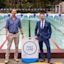 Two men stand beside a “Fast Lane” swim sign at an outdoor pool, one wearing a blue shirt and khakis, the other in a blue suit and tie.