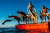 A group of people in swimwear jump and dive off a red floating platform into the water under a clear blue sky.