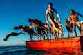 A group of people in swimwear jump and dive off a red floating platform into the water under a clear blue sky.