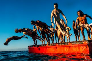 A group of people in swimwear jump and dive off a red floating platform into the water under a clear blue sky.
