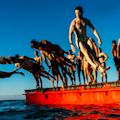 A group of people in swimwear jump and dive off a red floating platform into the water under a clear blue sky.