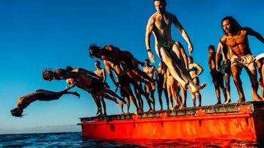 A group of people in swimwear jump and dive off a red floating platform into the water under a clear blue sky.