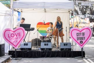 A small band performs on a stage with a rainbow heart backdrop and pink heart signs reading "LOVE SYDNEY XX" and "CITY OF SYDNEY" in an outdoor setting.