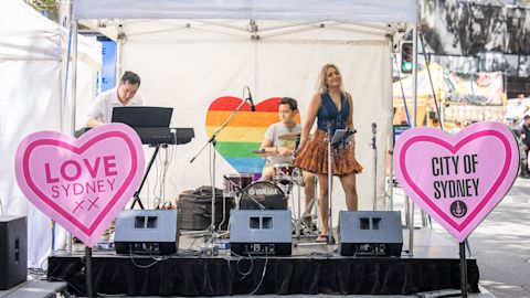 A small band performs on a stage with a rainbow heart backdrop and pink heart signs reading "LOVE SYDNEY XX" and "CITY OF SYDNEY" in an outdoor setting.