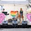 A small band performs on a stage with a rainbow heart backdrop and pink heart signs reading "LOVE SYDNEY XX" and "CITY OF SYDNEY" in an outdoor setting.