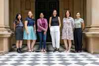 Six women stand in a row on a chequered floor in front of a building with columns and a large dark door.