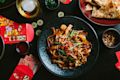 A black plate of stir-fried noodles with vegetables and seafood sits on a dark table surrounded by red envelopes, coins, flatbread, and drinks.