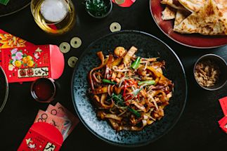 A black plate of stir-fried noodles with vegetables and seafood sits on a dark table surrounded by red envelopes, coins, flatbread, and drinks.