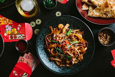 A black plate of stir-fried noodles with vegetables and seafood sits on a dark table surrounded by red envelopes, coins, flatbread, and drinks.