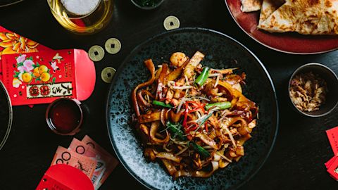 A black plate of stir-fried noodles with vegetables and seafood sits on a dark table surrounded by red envelopes, coins, flatbread, and drinks.