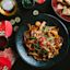 A black plate of stir-fried noodles with vegetables and seafood sits on a dark table surrounded by red envelopes, coins, flatbread, and drinks.