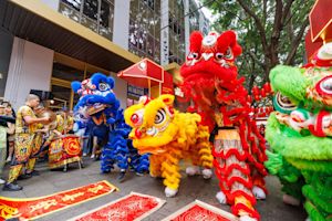 Performers in colorful lion costumes—red, green, yellow, and blue—dance on a city street during a festival, with onlookers and buildings in the background.