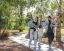 A family of five walks along a park path. The parents hold a baby and young children. Trees and greenery surround them in the background.