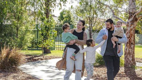 A family of five walks along a park path. The parents hold a baby and young children. Trees and greenery surround them in the background.