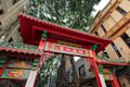 Red and green traditional Chinese gate with gold lettering stands between urban buildings and leafy trees.