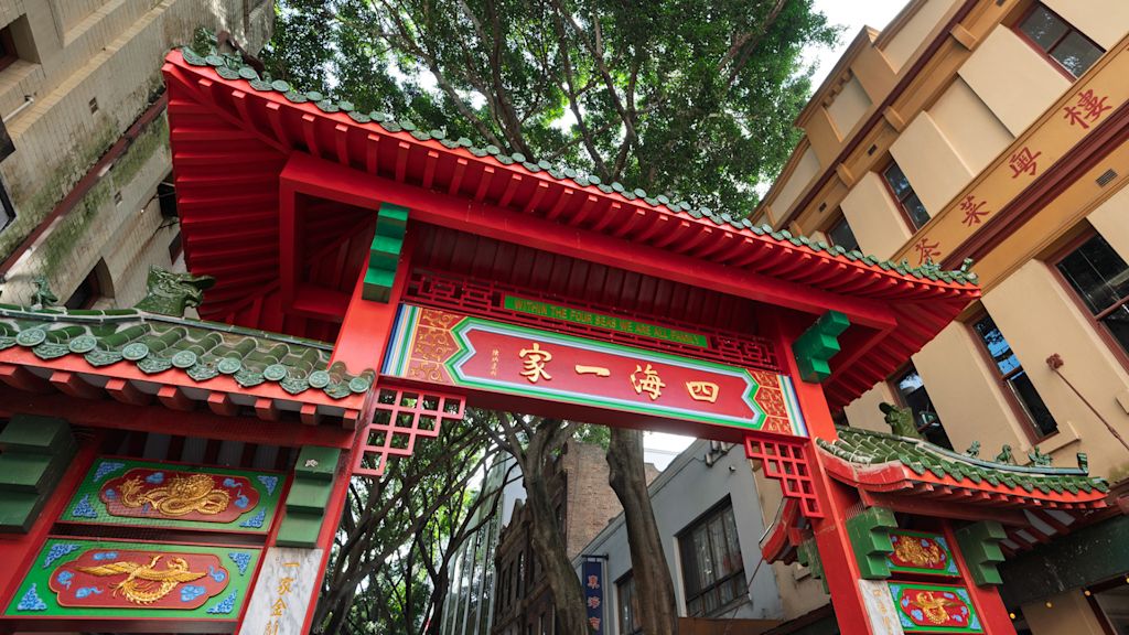Red and green traditional Chinese gate with gold lettering stands between urban buildings and leafy trees.