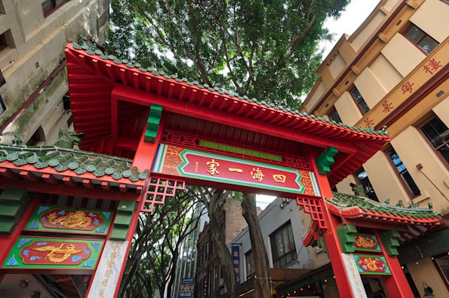 Red and green traditional Chinese gate with gold lettering stands between urban buildings and leafy trees.
