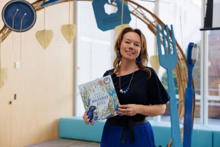 A woman holding a children's book titled "Bowerbird Blues" stands in front of a decorative arch with large blue utensils and hanging leaf decorations.