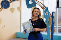 A woman holding a children's book titled "Bowerbird Blues" stands in front of a decorative arch with large blue utensils and hanging leaf decorations.