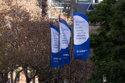 Three blue flags in front of trees with text that reads "Refugees and people seeking asylum are welcome in our city." The flags have the "City of Sydney" logo.